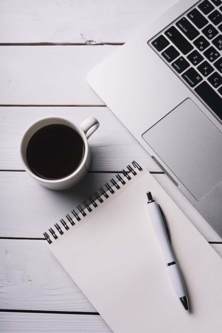 Coffee cup, notepad and pen, laptop on a desk.