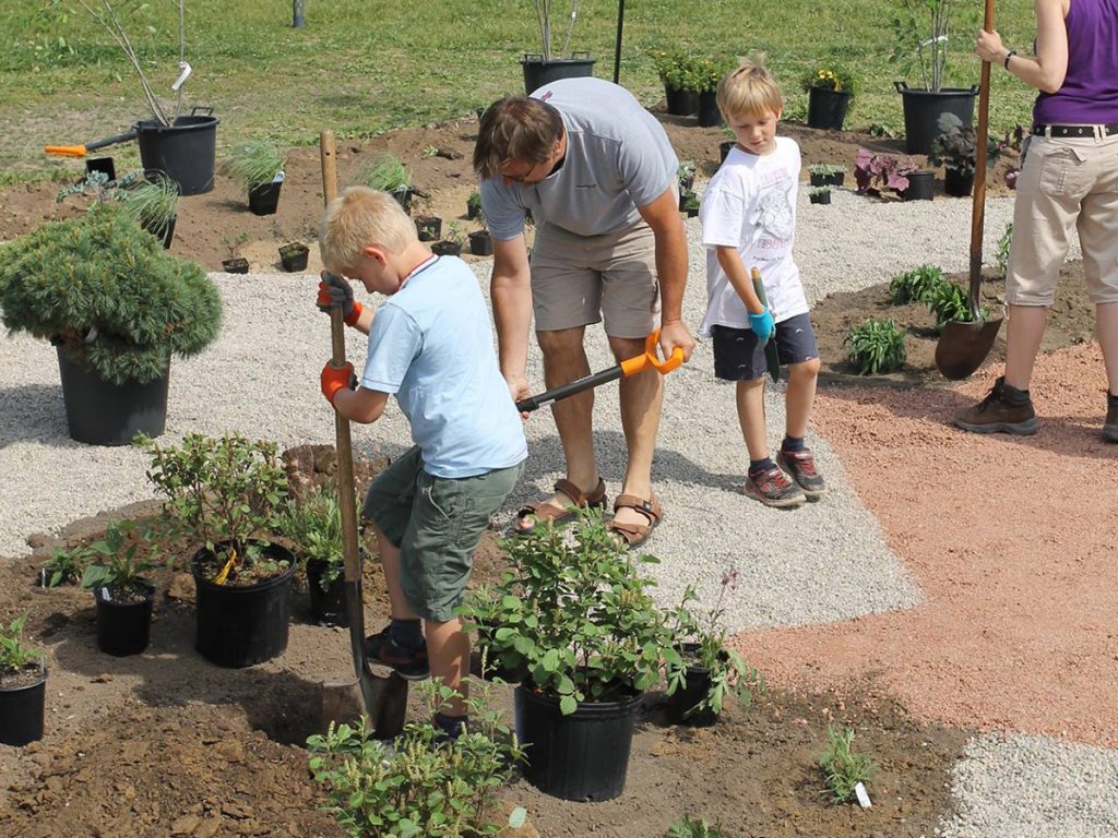 Two children and two adults work together, digging holes and planting greenery on a recently depaved asphalt playground. Potted plants surround them, with a school field in the background.