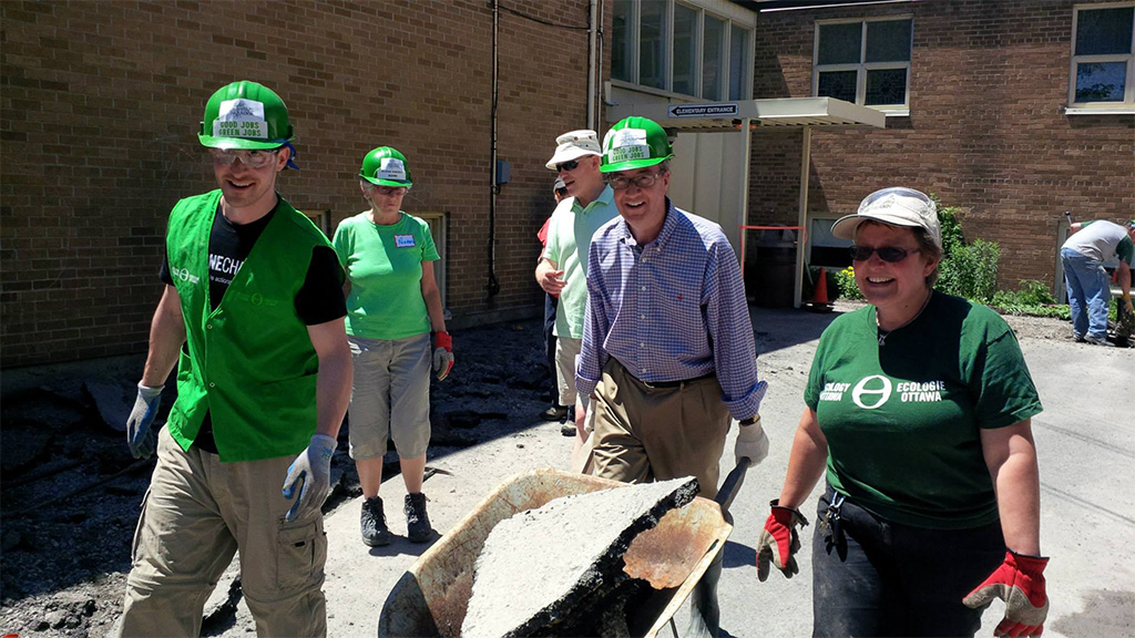 The mayor of Ottawa pushes a wheelbarrow filled with removed asphalt, with four smiling volunteers walking beside him.