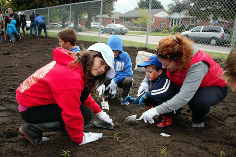 A group of children and an adult are crouched low over fresh laid soul, carefully tending to small plants with hand shovels