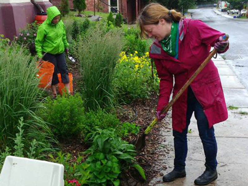 Two people tending to a roadside rain garden planted with shrubs and flowers. One carries buckets while the other digs into the soil with a shovel.