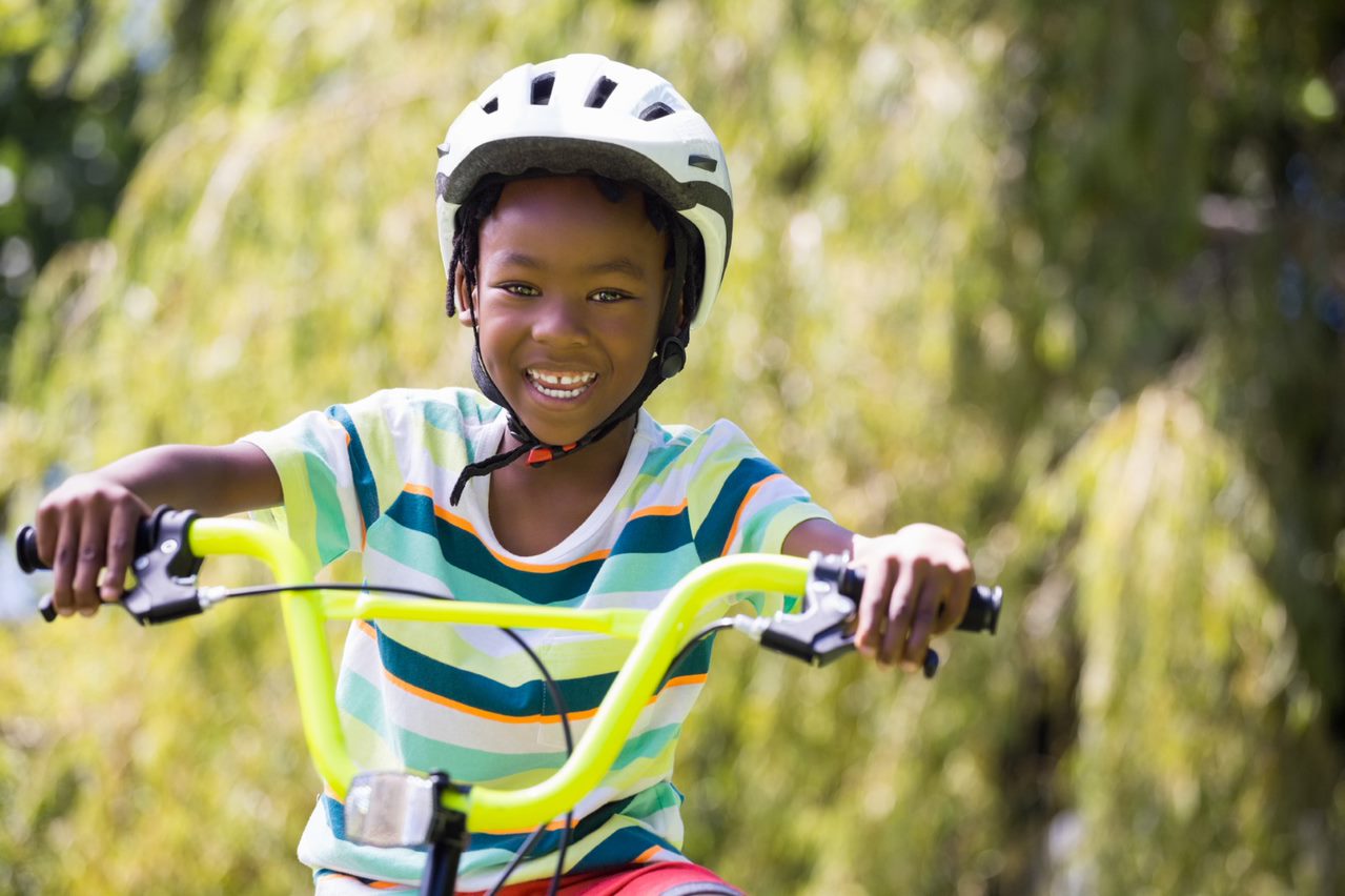 Child wearing a bike helmet smiles while riding a yellow bicycle outdoors.