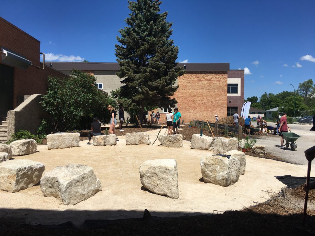 Outdoor classroom with large stone seats arranged in a circle. Students and volunteers work nearby, with the school building in the background.
