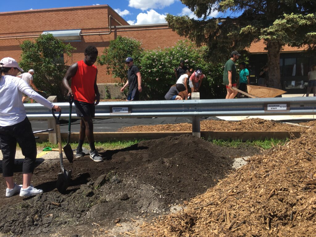 Students and volunteers shovel soil and move mulch. A wheelbarrow, tools, and a metal guardrail are visible in front of the school building.