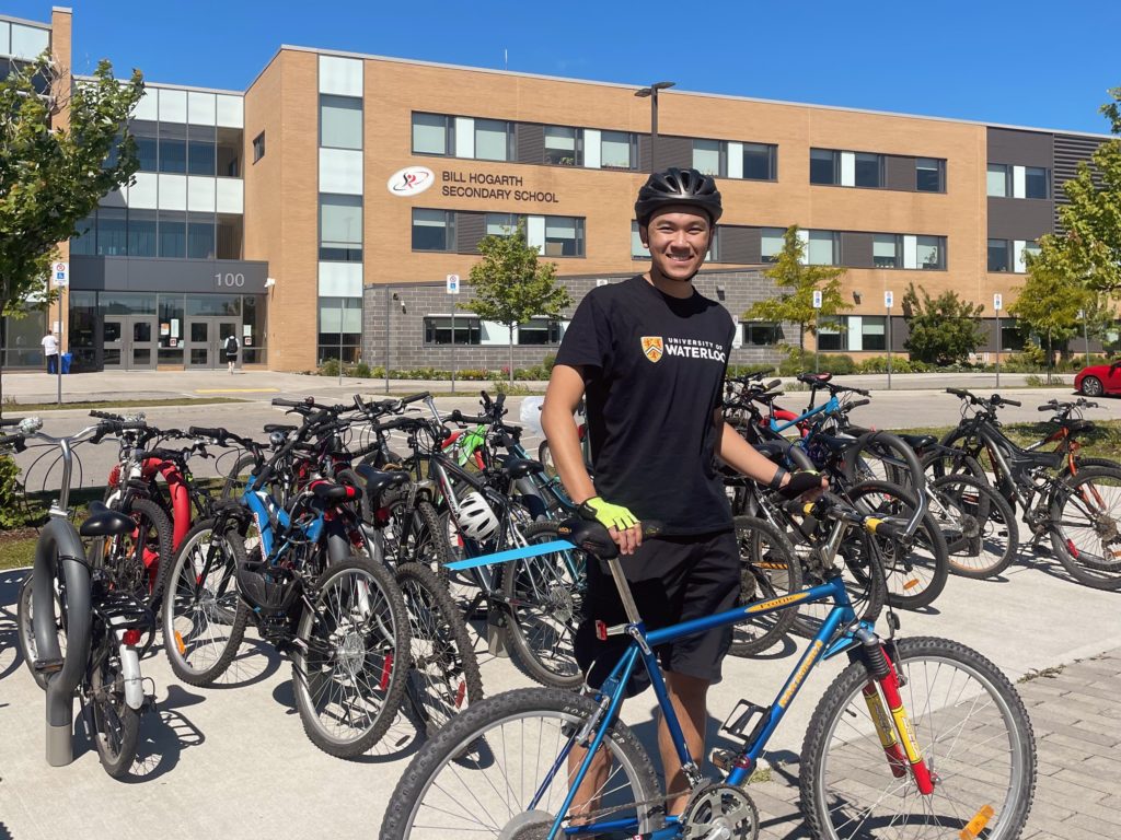Student wearing a helmet stands with a bicycle in front of a school building.