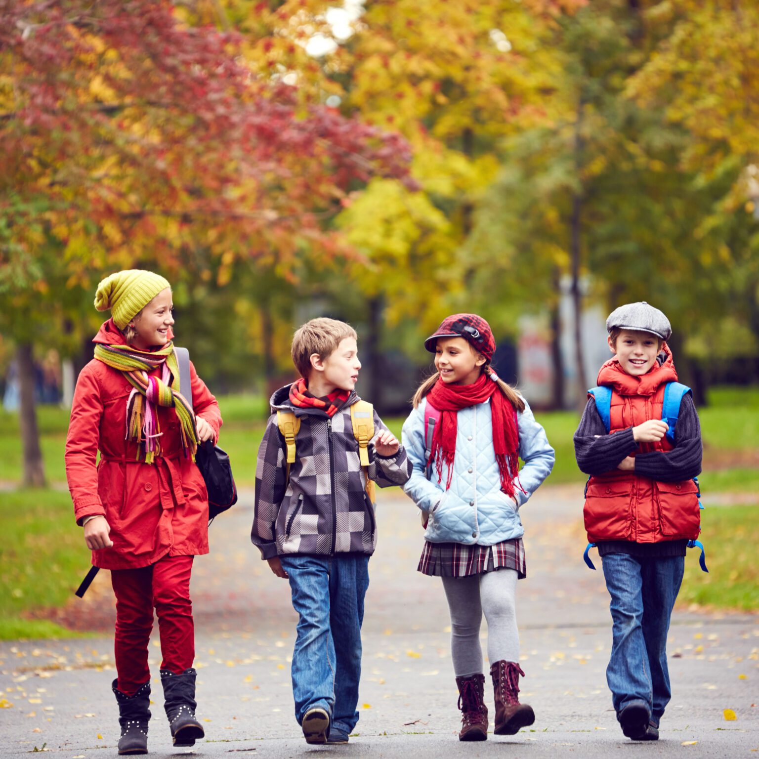 Four children walk together along a park path in autumn, dressed in colorful jackets, hats, and scarves.