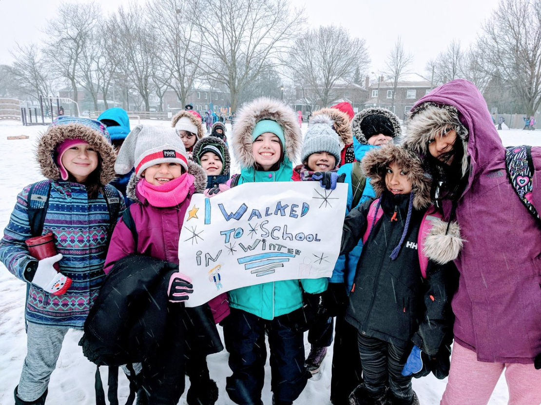 Group of children in winter coats and hats holding a handmade sign that reads “I walked to school in winter.”