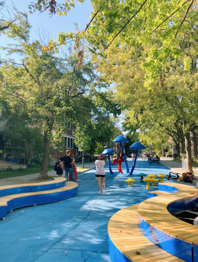 People walk on a tree-lined School Street with bright blue painted pavement, curved wooden seating, and large blue mushroom-shaped play structures.