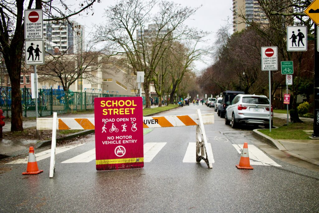 Barricades, traffic cones, and a bright sign reading “School Street - Road open to pedestrians, cyclists, and wheelchairs - No motor vehicle entry” block vehicle access to a city street outside a school.