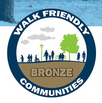 Walk Friendly Communities logo with a dark blue circle with the words "Walk Friendly Communities" around the edges. The centre of the circle has the word "Bronze" and a depiction of people holding hands and walking down a path.