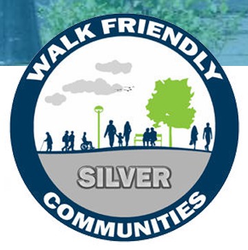 Walk Friendly Communities logo with a dark blue circle with the words "Walk Friendly Communities" around the edges. The centre of the circle has the word "silver" and a depiction of people holding hands and walking down a path.