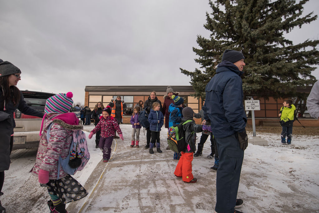 Children in winter clothing arrive at school on a snowy day, with parents and staff nearby supervising drop-off.