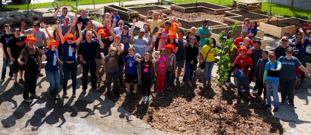 Smiling volunteers of all ages stand in a schoolyard with raised garden beds and a newly planted tree, waving at the camera after completing a depave project.